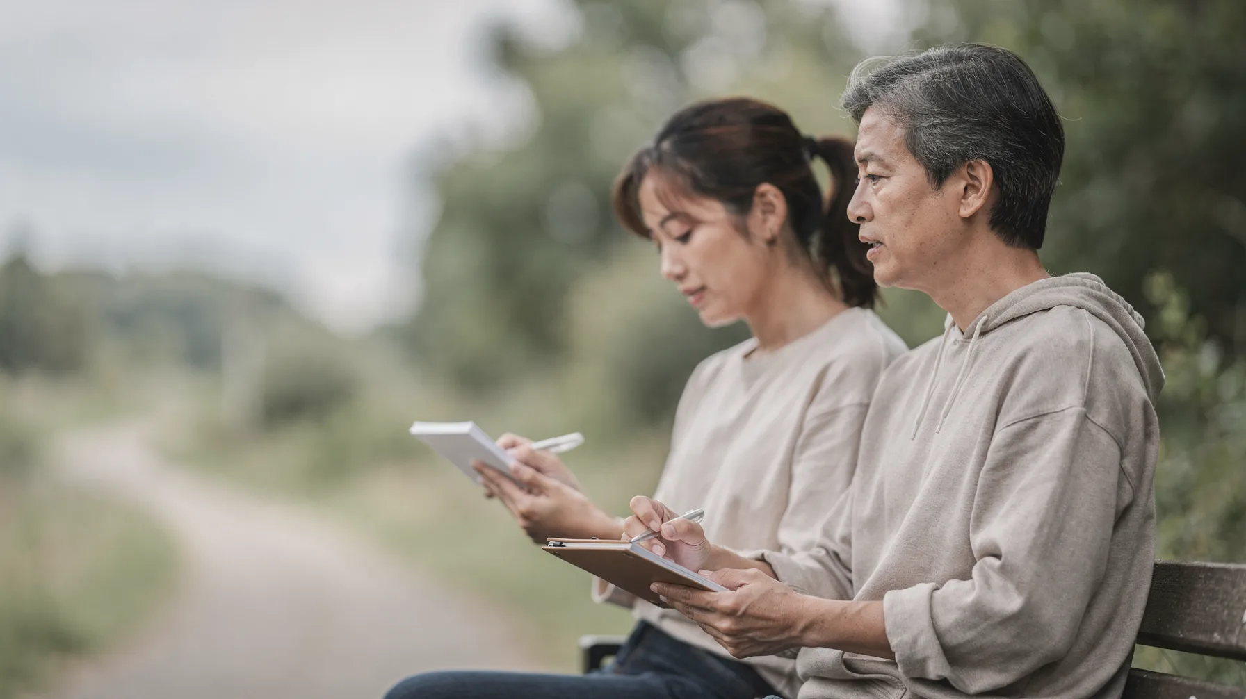 Two people seated on a park bench holding notebooks and pens.